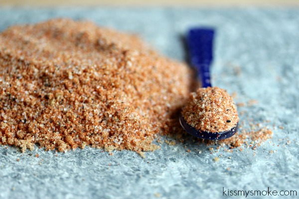 Smoked paprika rub is scattered on a blue-grey counter with a blue spoon on it that has the spice inside of it.