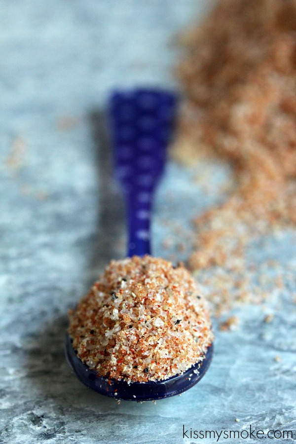 Smoked paprika rub is scattered on a blue-grey counter with a blue spoon on it that has the spice inside of it.