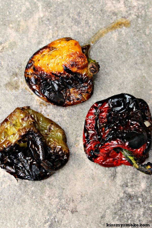Fire Roasted Bell Peppers on a grey surface, one red, one green and one yellow pepper are all charred and ready to have skins removed and insides scooped out.