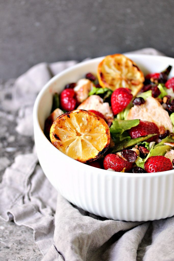 Grilled chicken salad with strawberries served in a large white bowl with grilled lemon halves for drizzling over top.