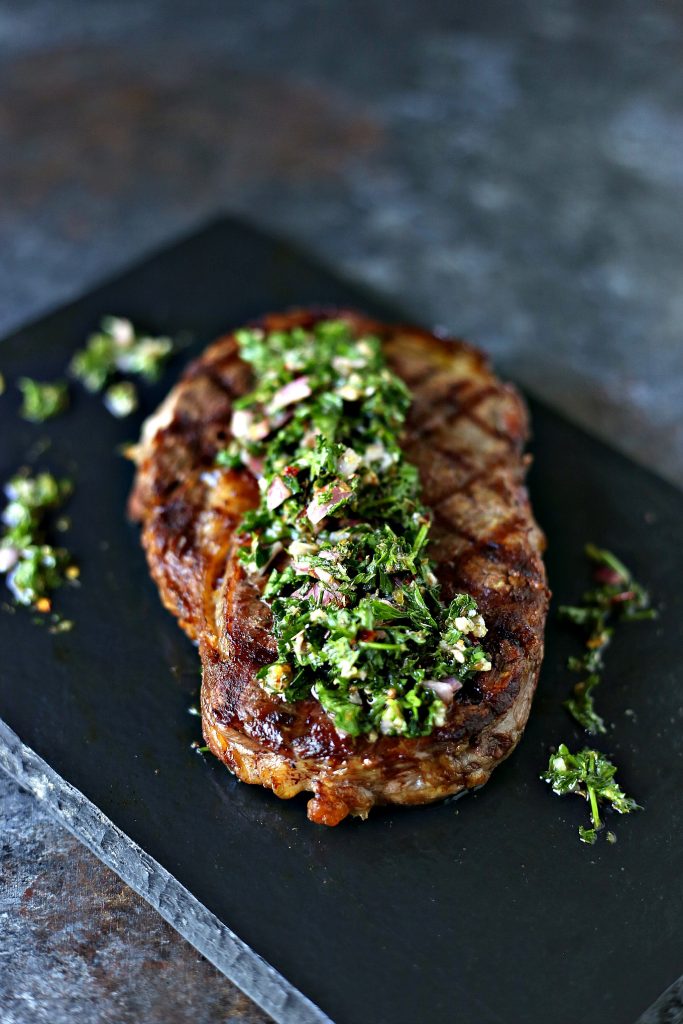 Grilled Rib Steaks with Chimichurri Sauce with perfect grill marks set on a black serving tray on a dark counter. 