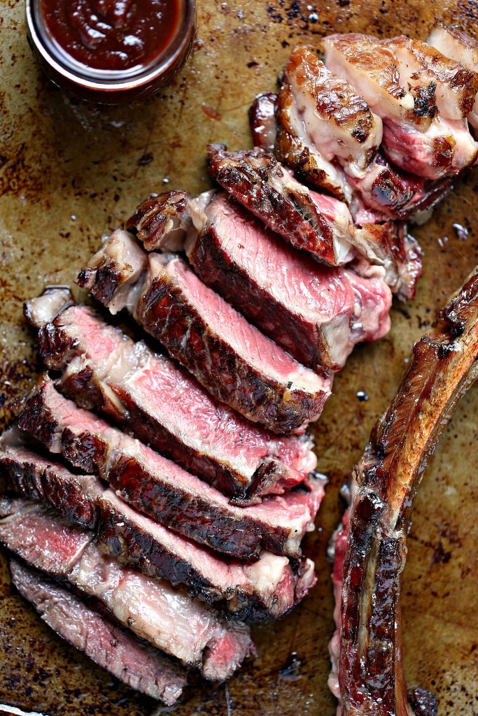 Overhead image of a grilled tomahawk steak that has been cooked and sliced. Bone has been cut off and is set next to the steak on the right side. A mason jar of bbq sauce is in the upper left corner.