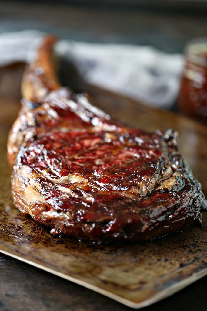 Portrait image of a grilled tomahawk steak resting after it was cooked. Steak is sitting on a metal sheet pan with a grey napkin and a small mason jar filled with bbq sauce in the background.
